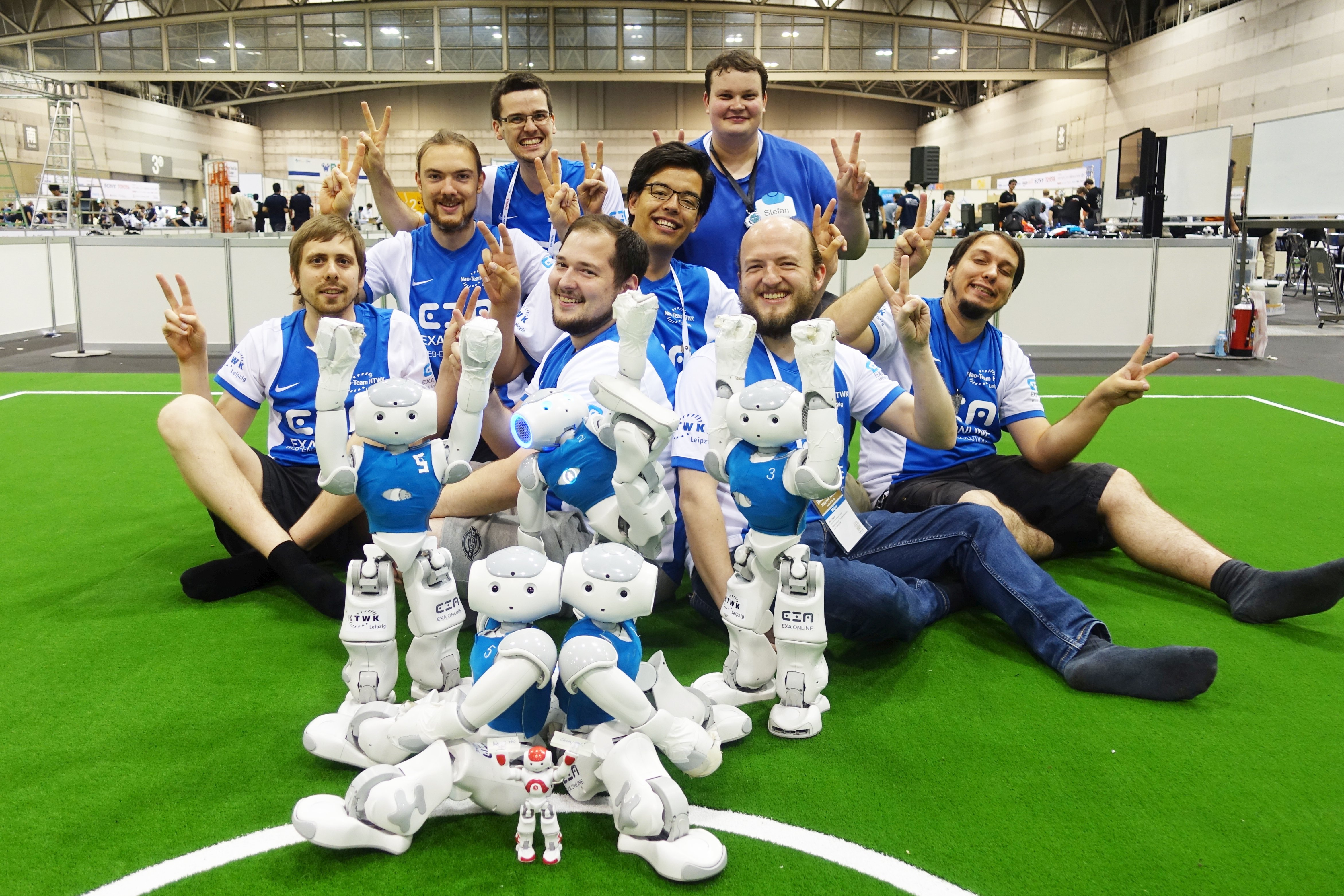 Gruppenfoto einen 8-köpfigen Teams lachend mit kleinen humanoiden Robotern vor sich auf dem Fußballspielfeld.