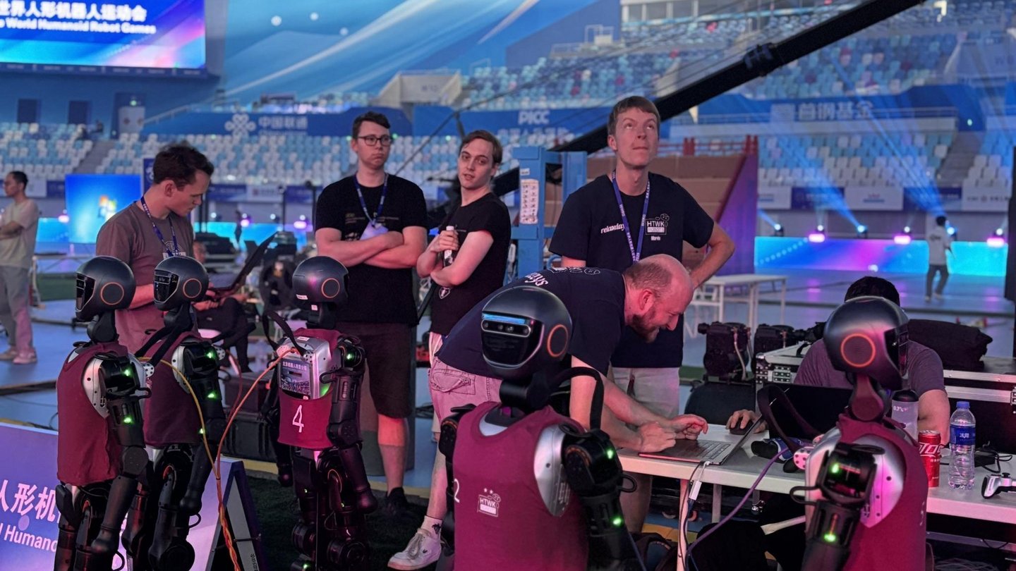 Mehrere Menschen und kindergroße humanoide Roboter zusammen in einem leerem Stadion beim Technikcheck.