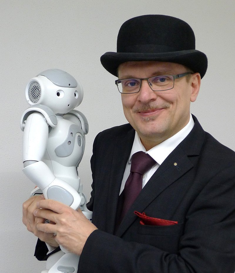 Lächelnder Mann im Anzug und mit Hut hält einen kleinen humanoiden Roboter in den Armen, der ihn anschaut.