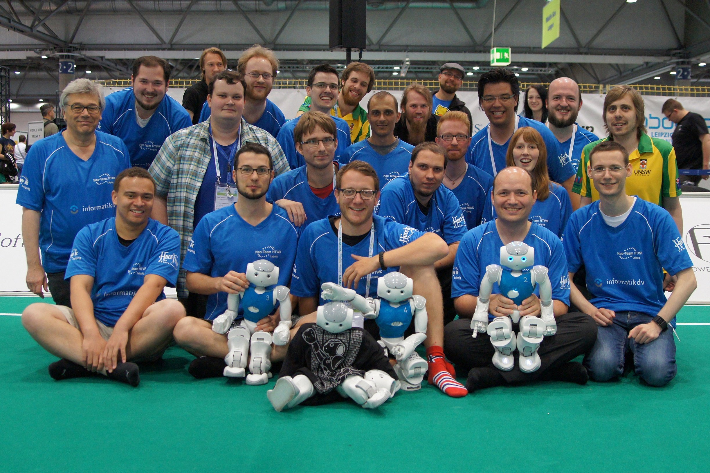 Gruppenfoto einen 21-köpfigen Teams lächelnd mit kleinen humanoiden Robotern vor sich auf dem Fußballspielfeld.