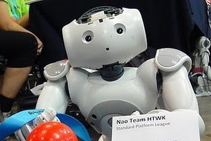Kleiner humanoider Roboter sitzt an einem kleinen Tisch, vor ihm ein Fußball und ein Schild mit Aufschrift des HTWK-Nao-Teams.