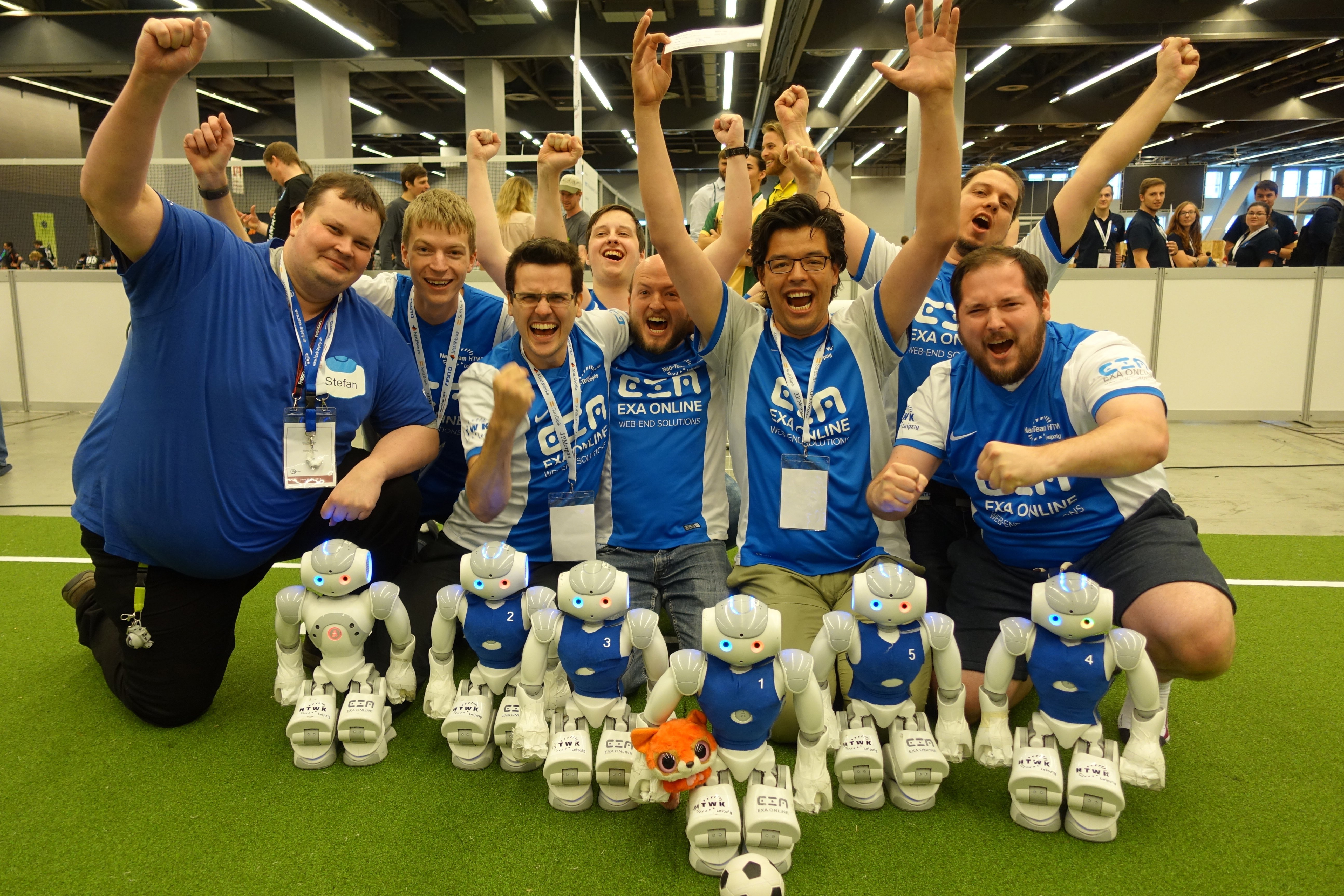 Gruppenfoto einen 8-köpfigen Teams jubeld mit kleinen humanoiden Robotern vor sich auf dem Fußballspielfeld.