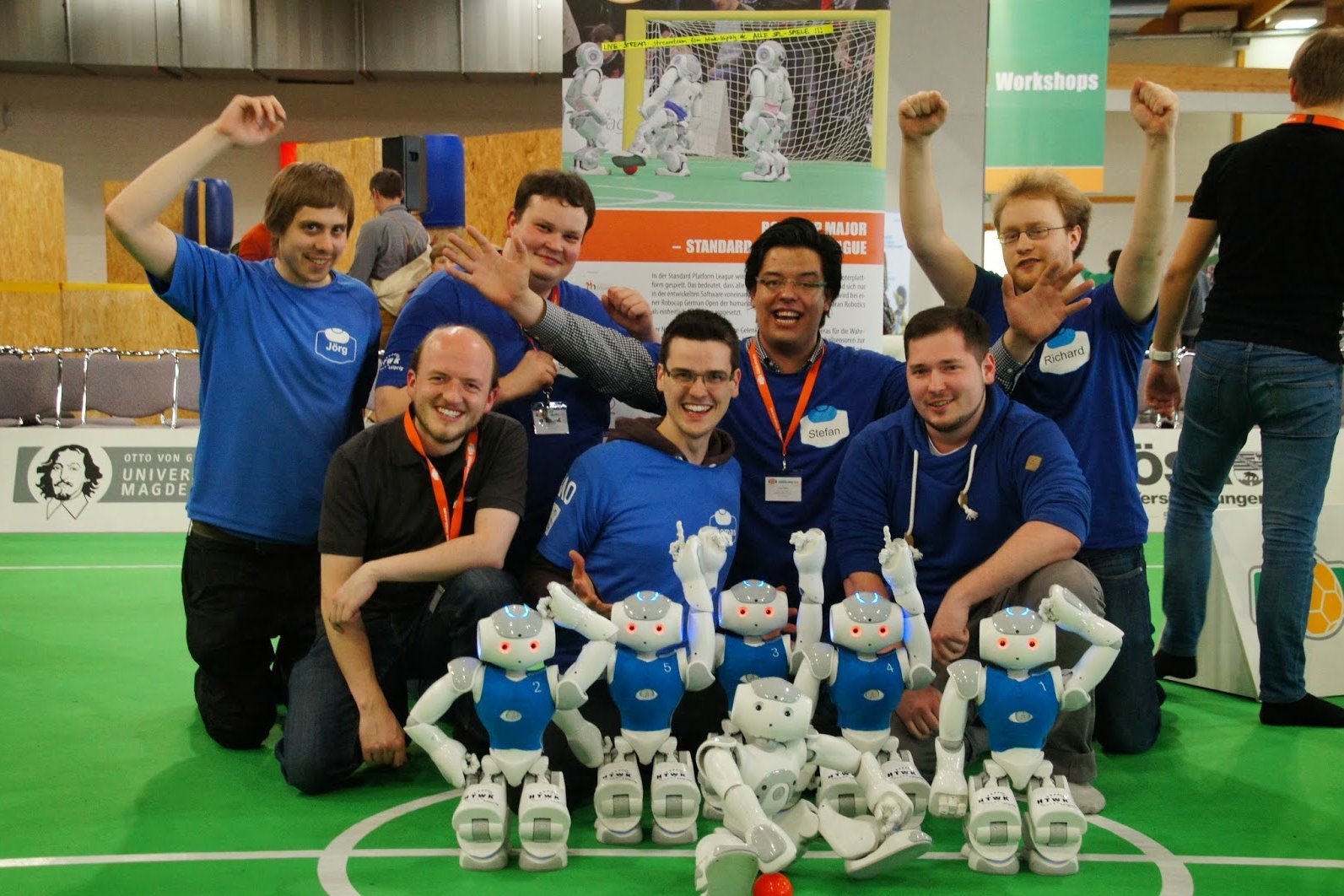 Gruppenfoto einen 7-köpfigen Teams jubeld mit kleinen humanoiden Robotern vor sich auf dem Fußballspielfeld.