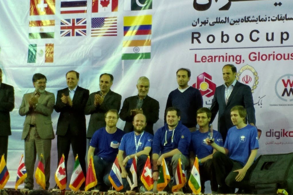 Gruppenfoto eines 5-köpfigen Teams kniend auf einer Bühne aufgereiht, hinter ihnen sechs applaudierende Männer im Anzug aufgereiht vor einem Plakat mit verschiedenen Länderflaggen.