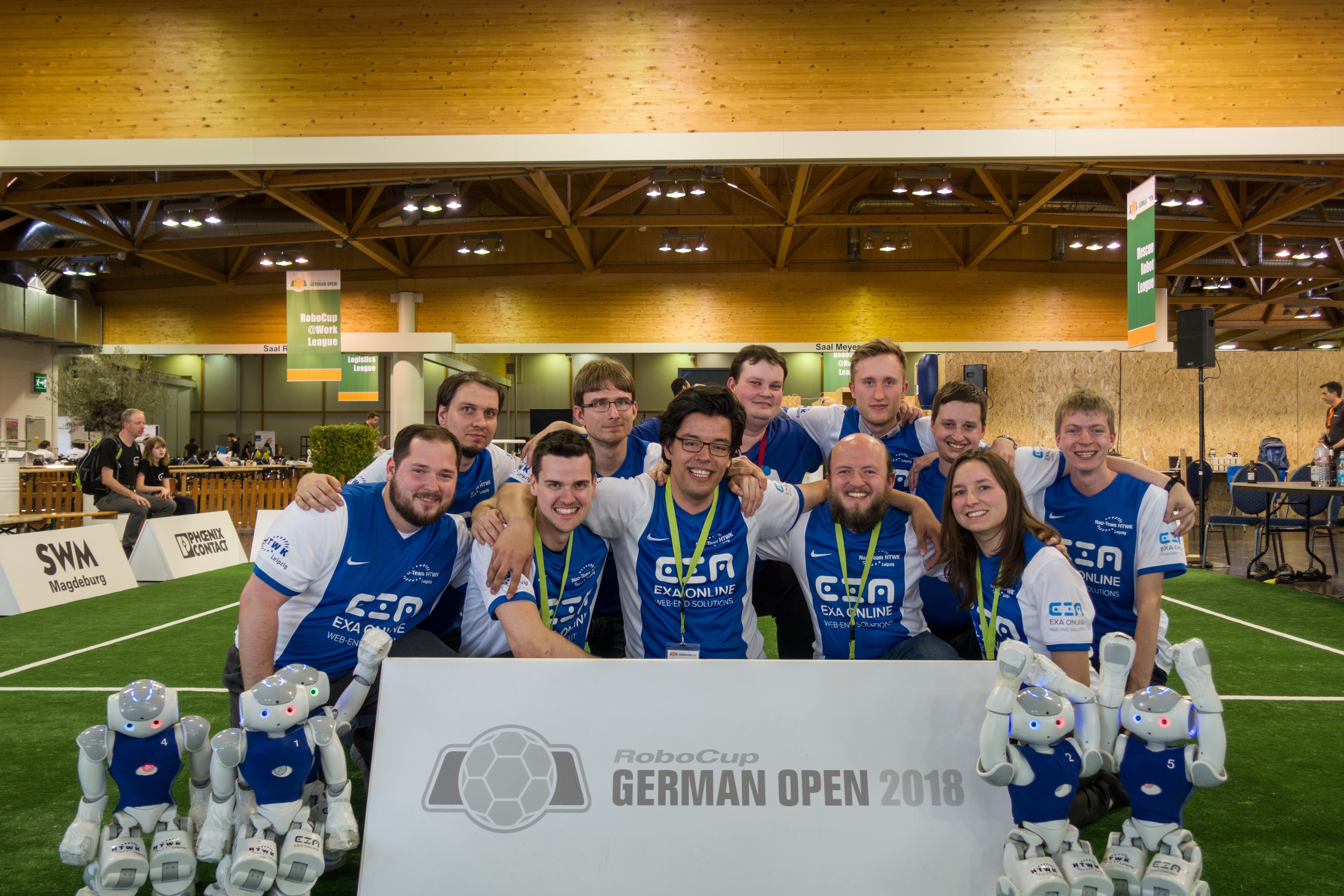 Gruppenfoto einen 11-köpfigen Teams mit Armen ineinander eingehakt und mit kleinen humanoiden Robotern vor sich auf dem Fußballspielfeld.