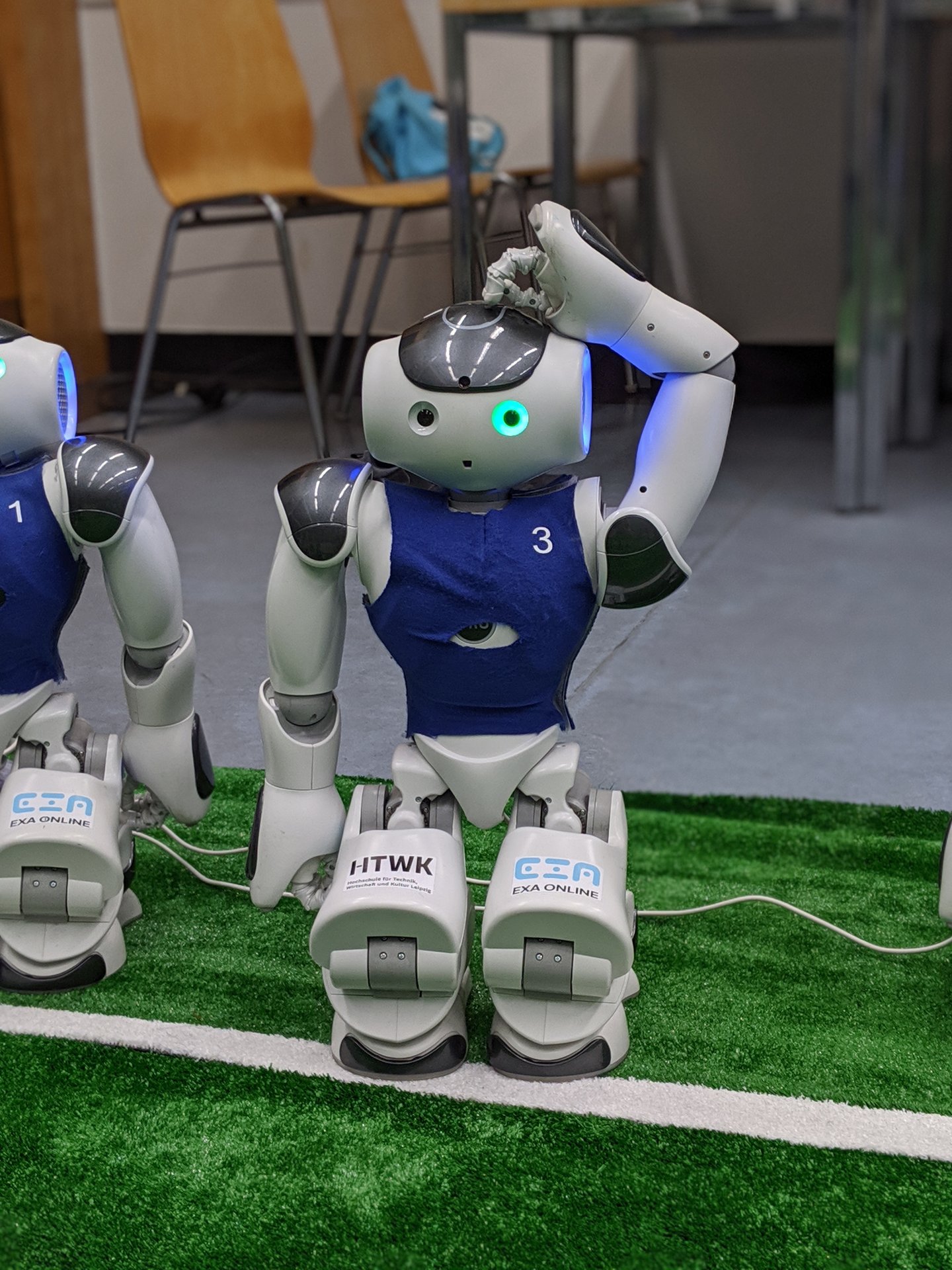Kleiner humanoider Roboter an der Seitenlinie mit eingestecktem Ladekabel und fragendem Blick hält seine Hand an seinen Kopf, als würde er seinen Hinterkopf kratzen.