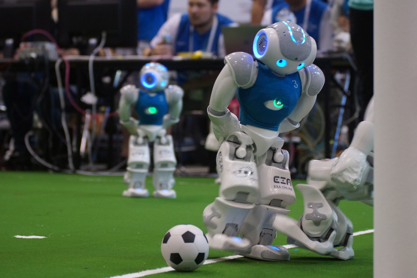 Zwei kleine humanoide Roboter auf einem Fußballfeld, wobei ein Roboter kurz vorm Umfallen ist. Im Hintergrund ein weiterer Mitspieler und am Seitenrand ein Publikum aus Menschen.